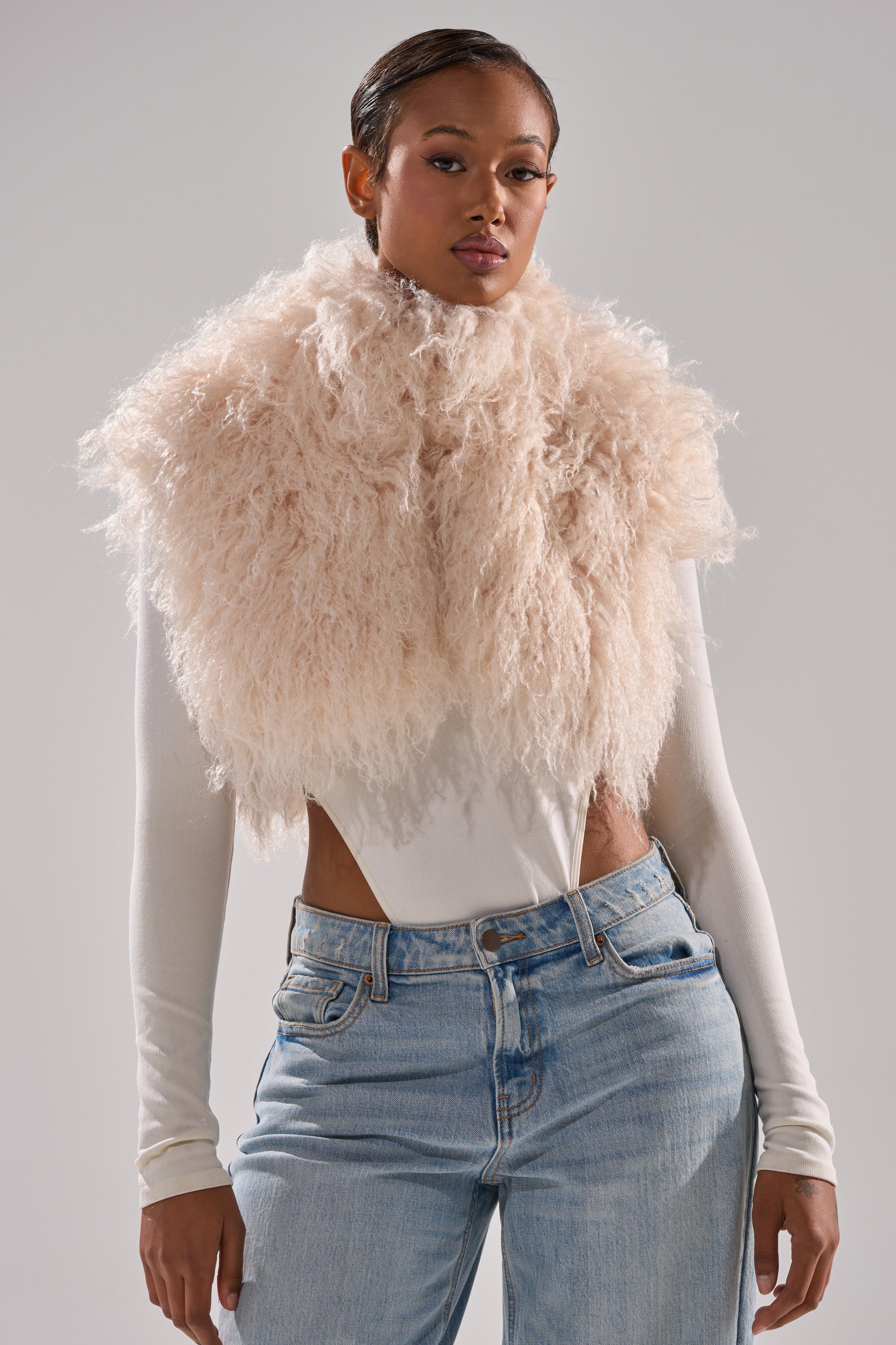A woman stands confidently against a plain background, wearing the COCO CROPPED FUR VEST IN BEIGE over a white long-sleeve bodysuit and light blue jeans.