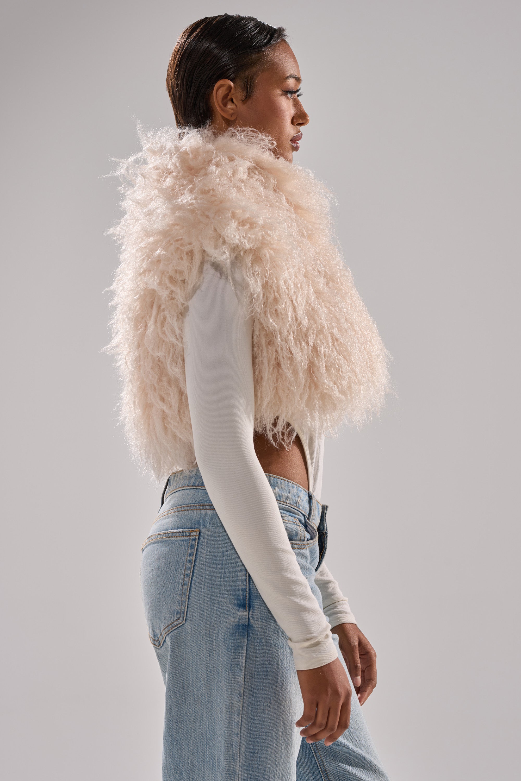 A person in profile wears the COCO CROPPED FUR VEST IN BEIGE over a white long-sleeve top and light blue jeans, with slicked-back hair against a plain light gray background.
