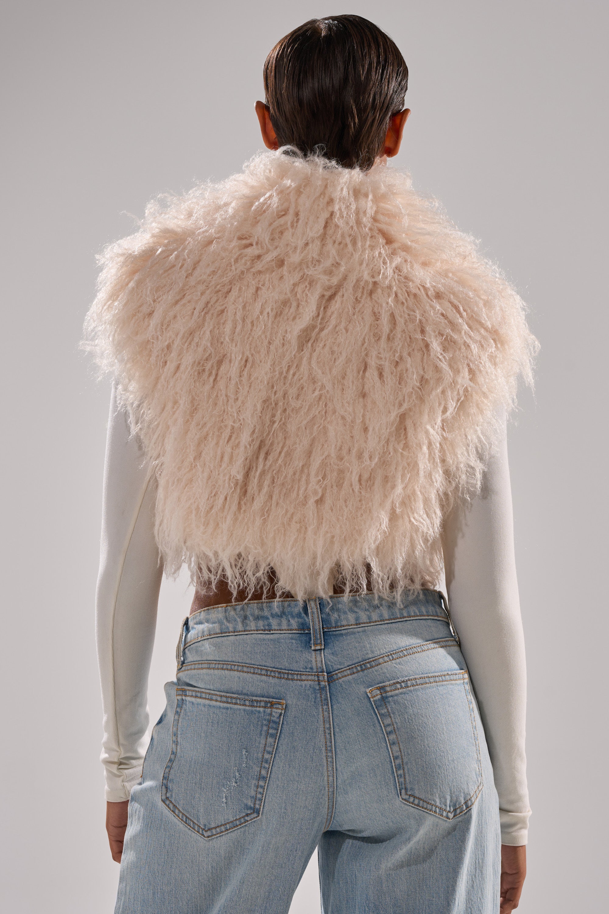 A person with slicked-back hair stands against a light gray background, wearing the COCO CROPPED FUR VEST IN BEIGE over a long-sleeved white top and light blue jeans.