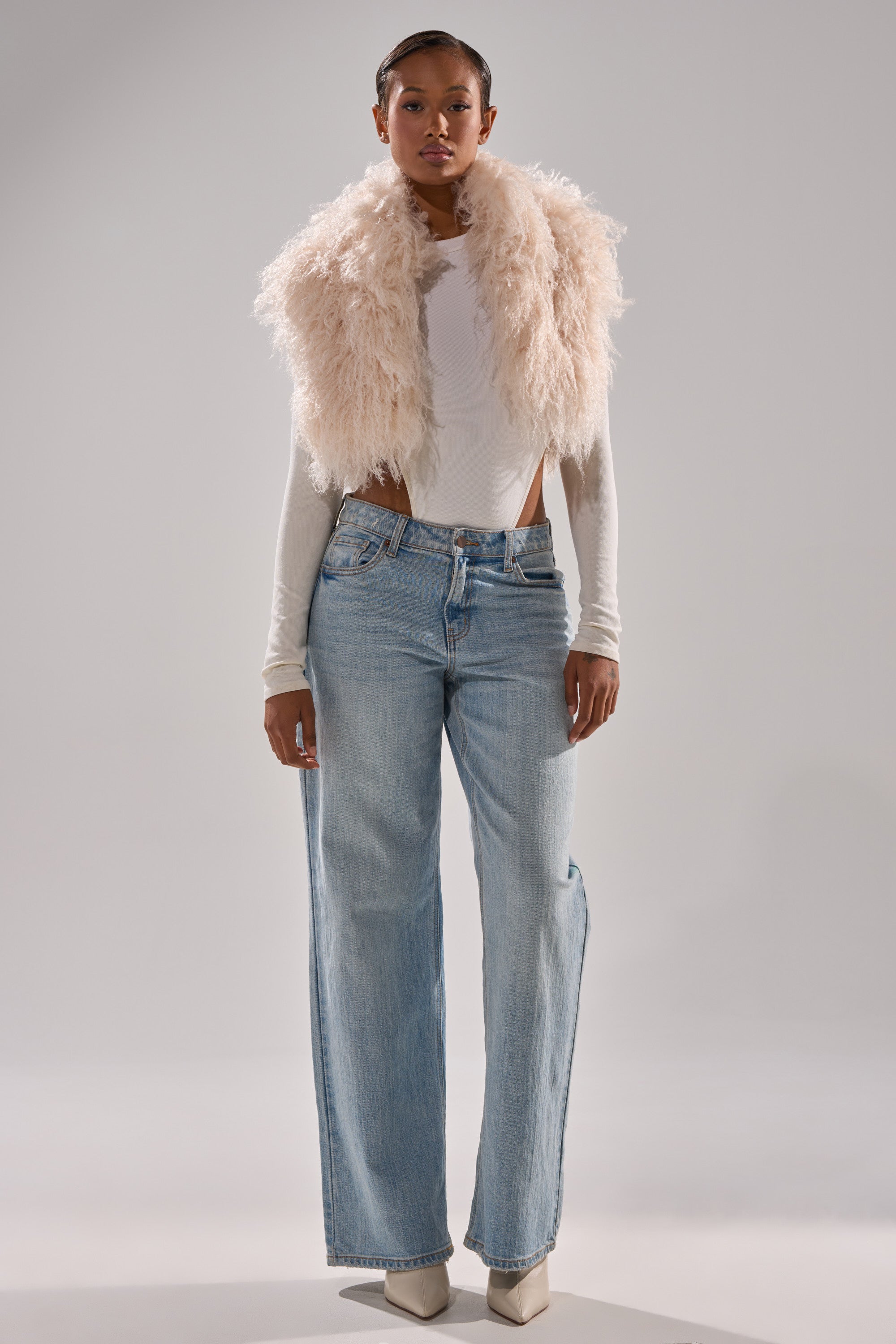 A person stands against a plain background wearing the COCO CROPPED FUR VEST IN BEIGE over a fitted white top, paired with light blue high-waisted wide-leg jeans and pointed white shoes.