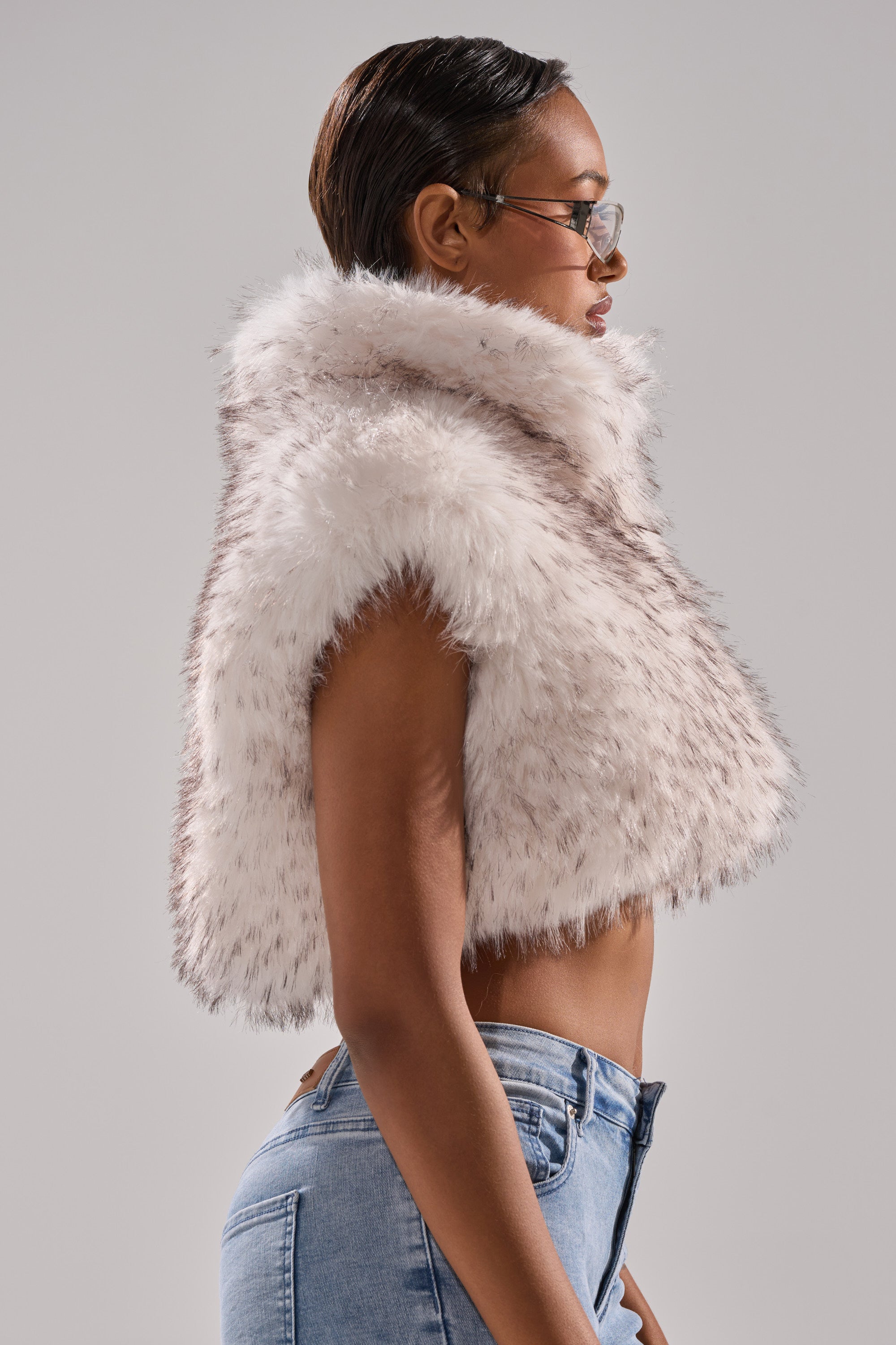 A person with short, slicked-back hair and glasses models the COCO CROPPED FUR VEST IN WHITE MULTI with blue jeans, posing in profile against a plain gray background.