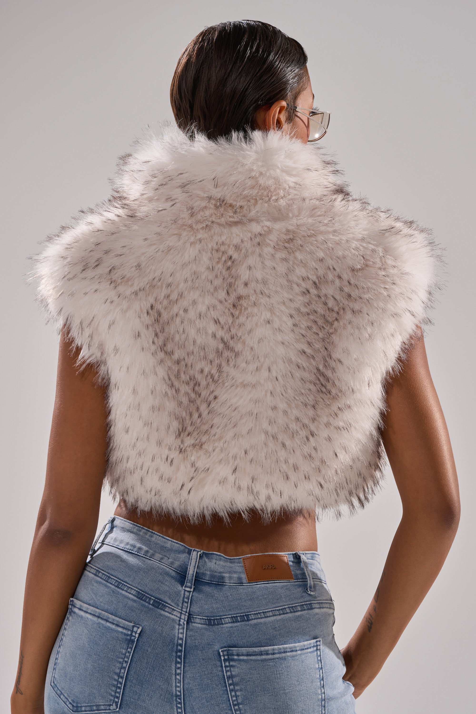 A person is seen from behind wearing the COCO Cropped Fur Vest in White Multi, featuring dark specks, with light blue jeans and slicked-back hair, standing against a plain gray background.