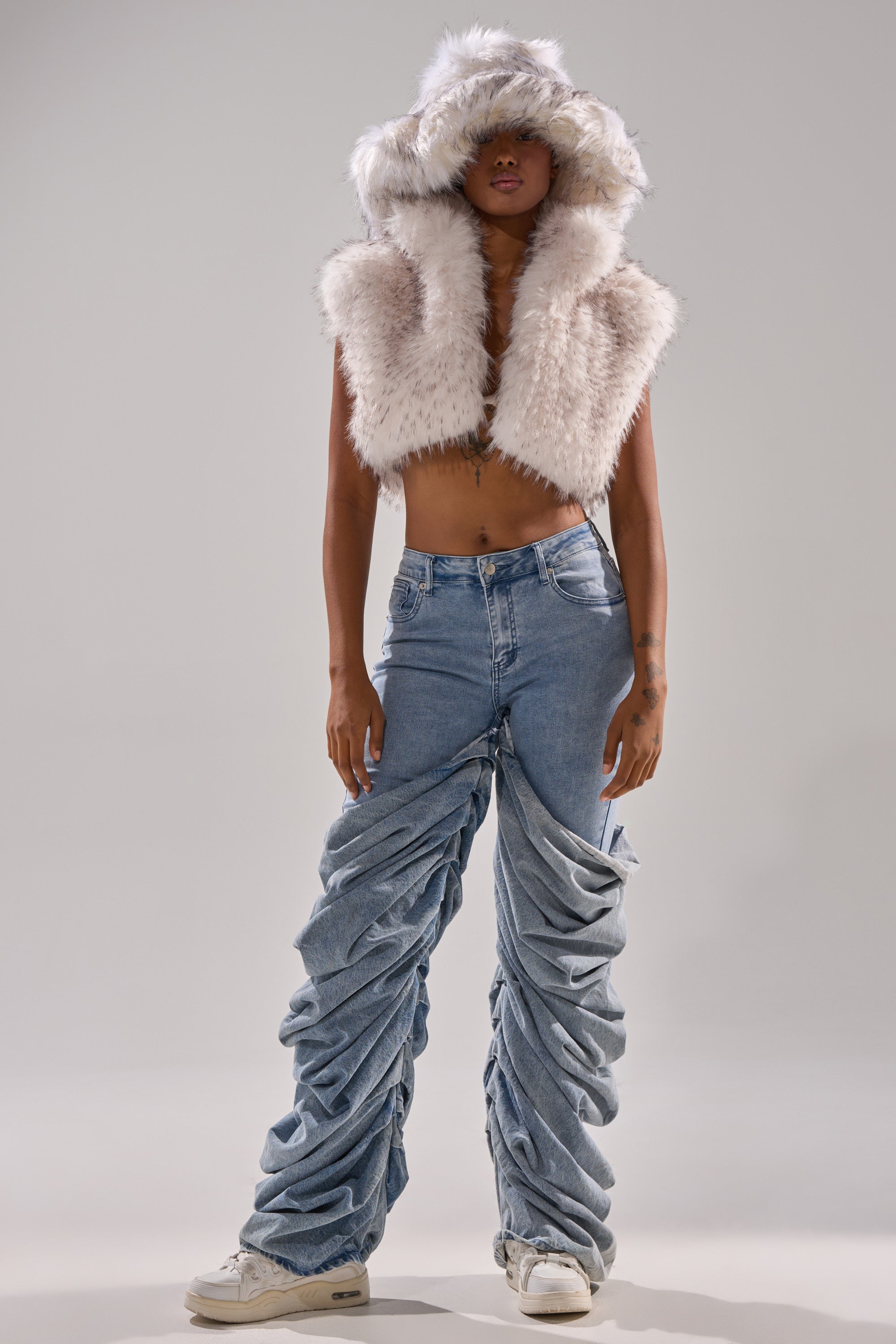 Wearing the COCO CROPPED FUR VEST IN WHITE MULTI with a fluffy hood partly covering their face, white sneakers, and low-rise ruched denim jeans, the person stands against a plain background with their midriff exposed.