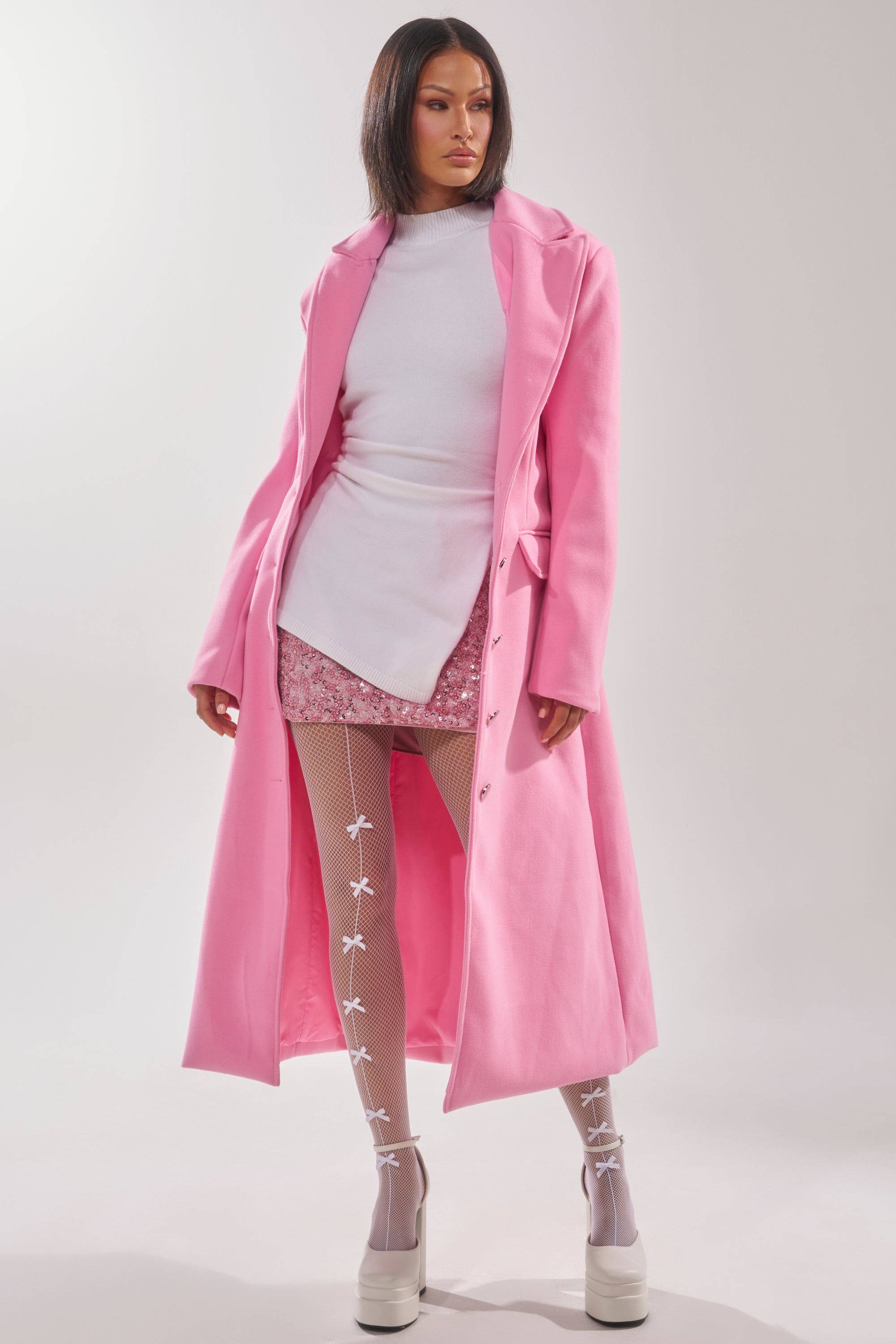 A woman stands confidently against a plain background wearing the SWEET THING WOVEN TRENCH, a pink trench coat styled with a white turtleneck, short pink patterned skirt, white bow tights, and platform shoes. 2