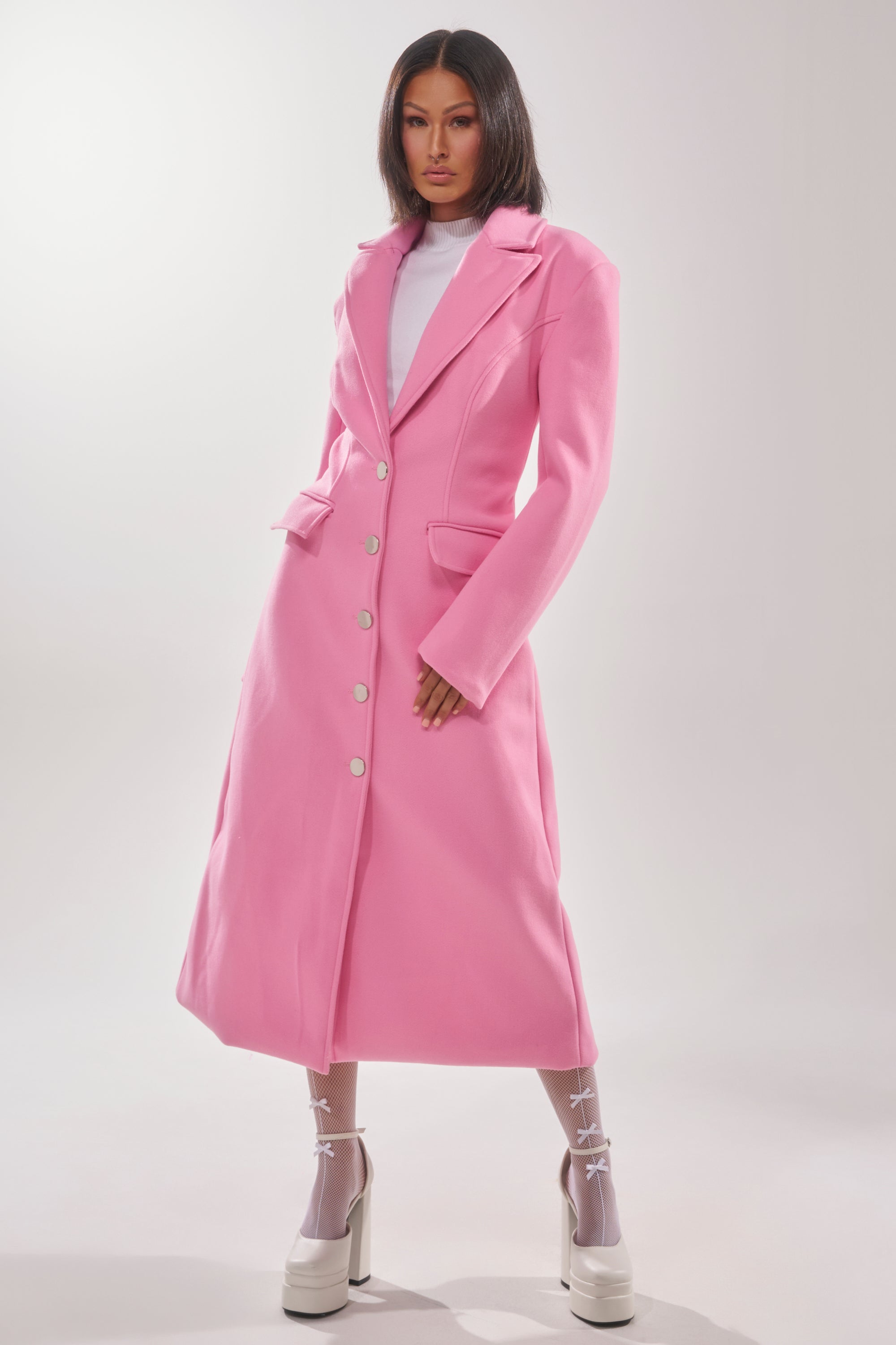A woman exudes confidence in the SWEET THING WOVEN TRENCH—a long, bright pink coat with white buttons over a white top and platform heels. The light background accentuates her stylish, modern look and bold statement outerwear. 3
