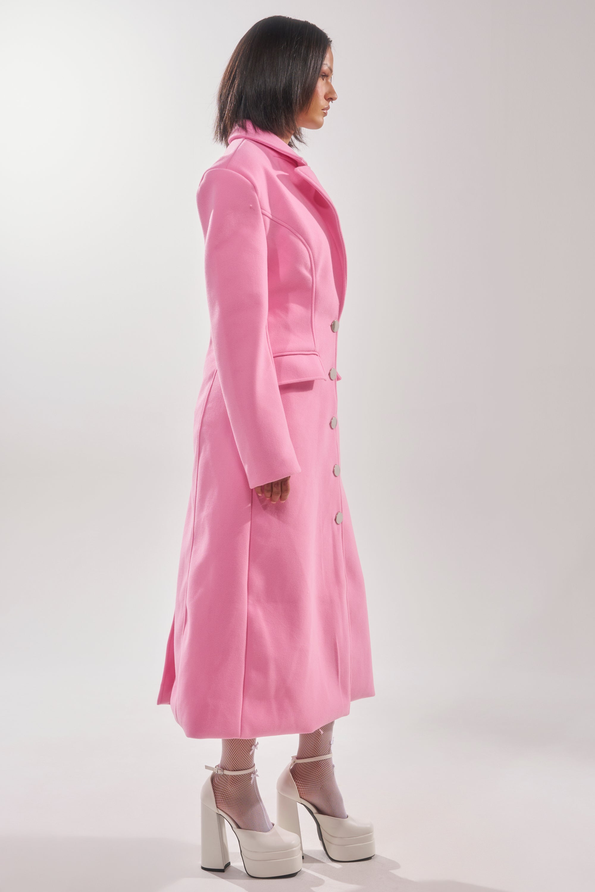 A person stands in profile against a plain, light background wearing the SWEET THING WOVEN TRENCH, paired with sheer socks and white platform shoes. 4