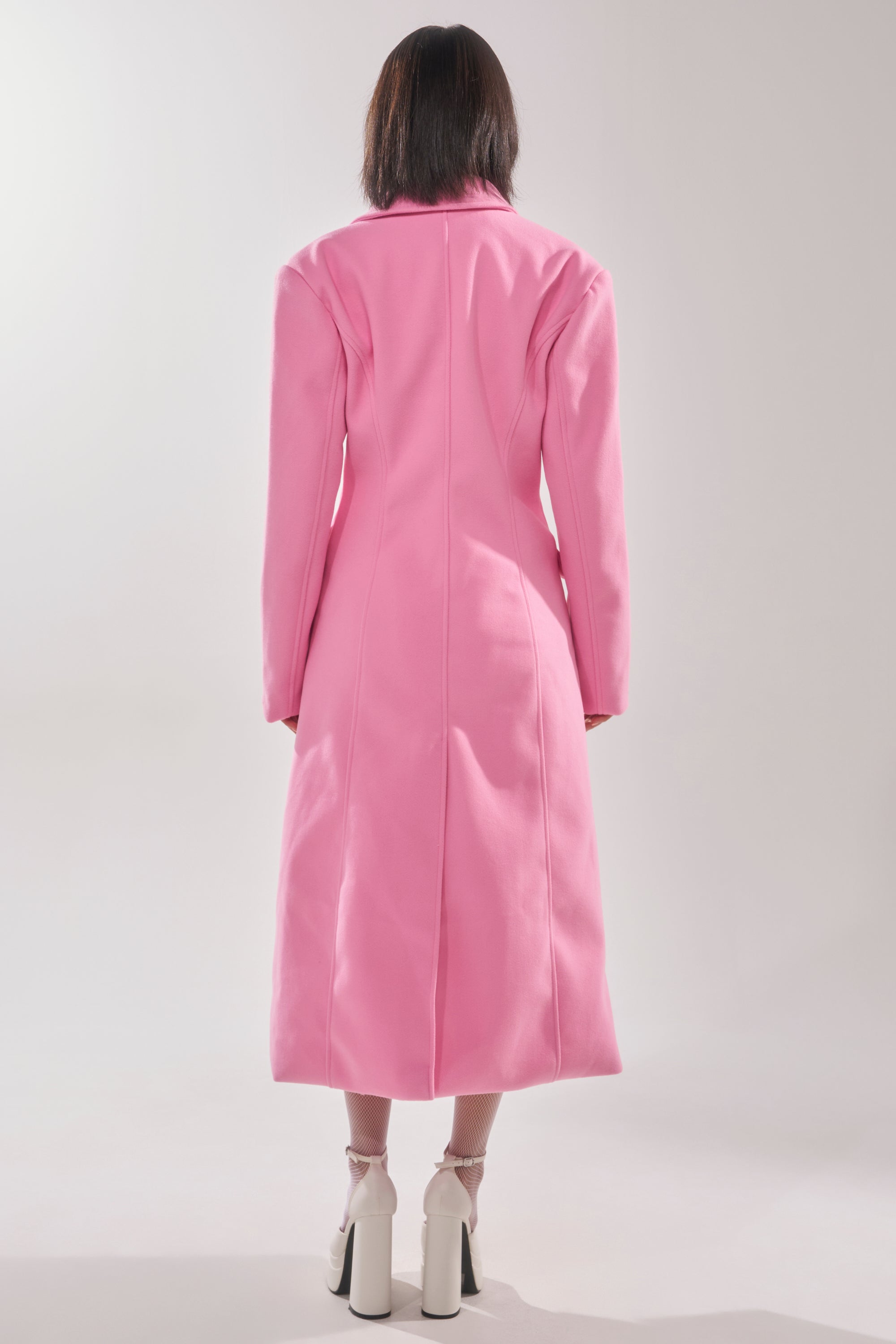 A person faces away, wearing the SWEET THING WOVEN TRENCH in bright pink as a statement jacket, paired with white ankle-strap high heels, all set against a neutral light gray background. 5
