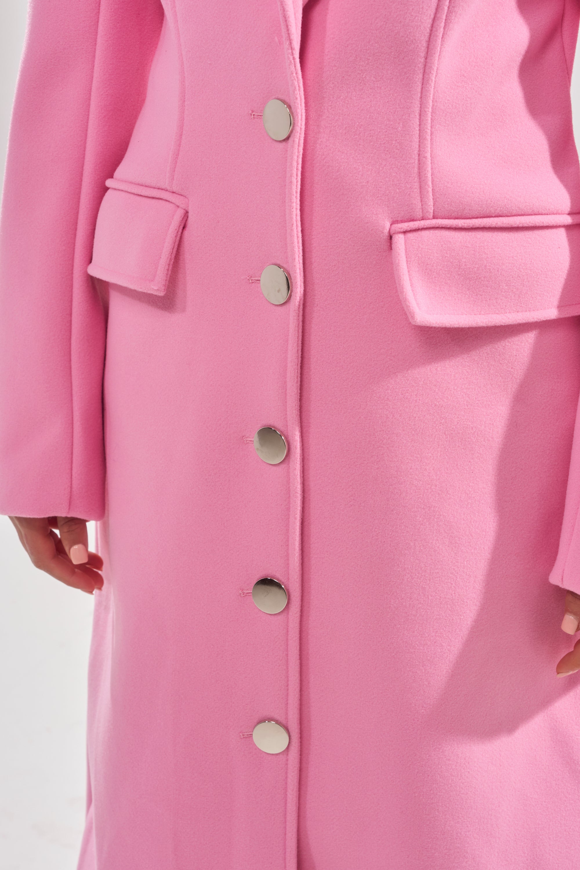 A close-up of a person wearing the SWEET THING WOVEN TRENCH, a long, bright pink coat with silver buttons and flap pockets, showcased from shoulders to knees. 7