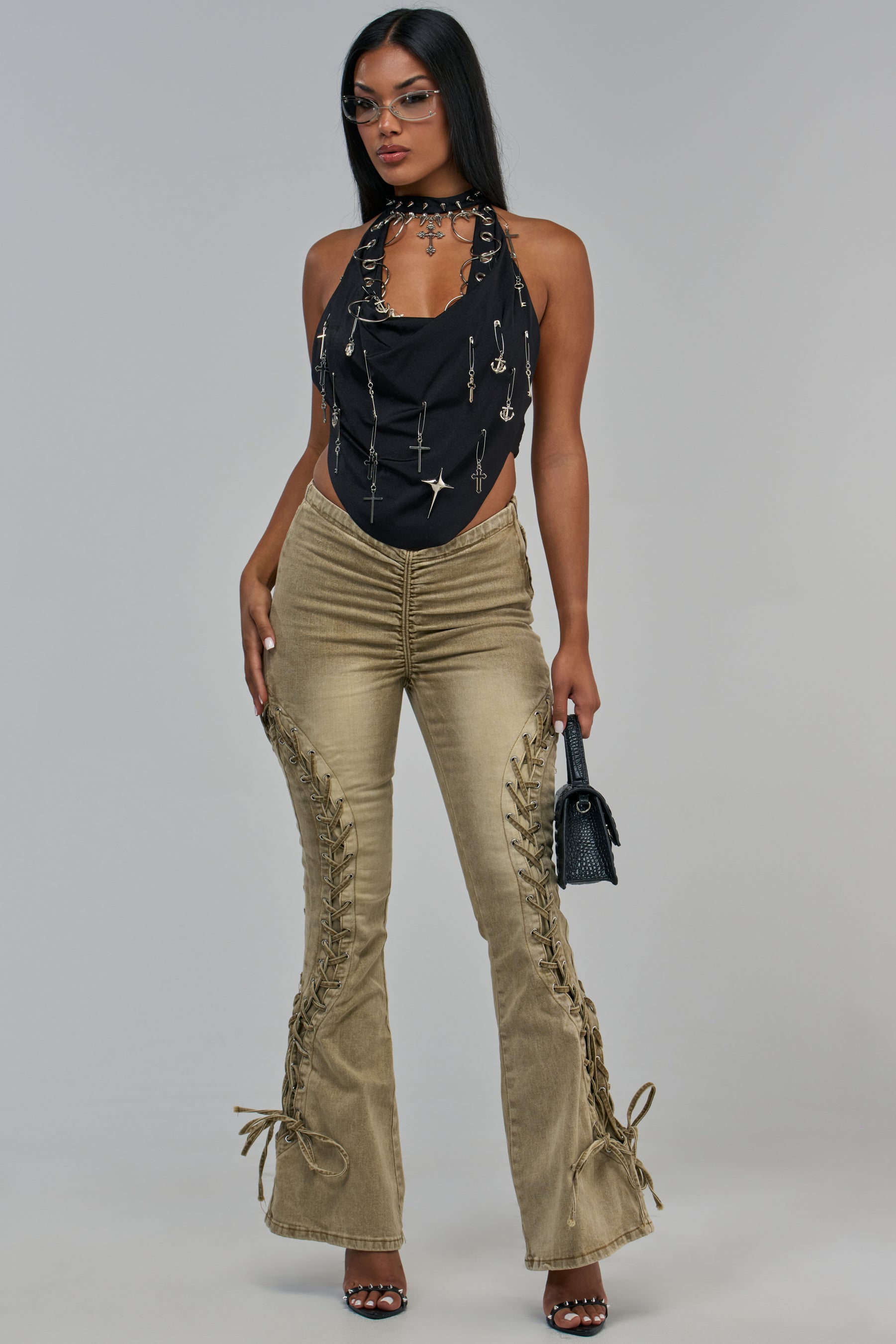 A woman wears a black halter top with silver charms and taupe TAKE NOTES LACE UP SCRUNCH DENIM PANTS featuring ruching at the waist and lace-up details along the outer legs.