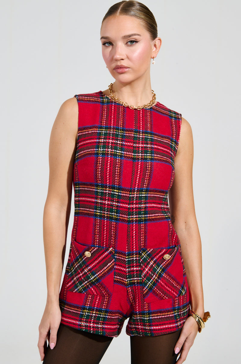 A person wearing the Best Dressed Plaid Mini Dress in red, featuring a sleeveless design with pockets and button details. The dress showcases a tweed-like fabrication, complemented by a gold chain necklace and matching bracelet against a plain, light-colored background.