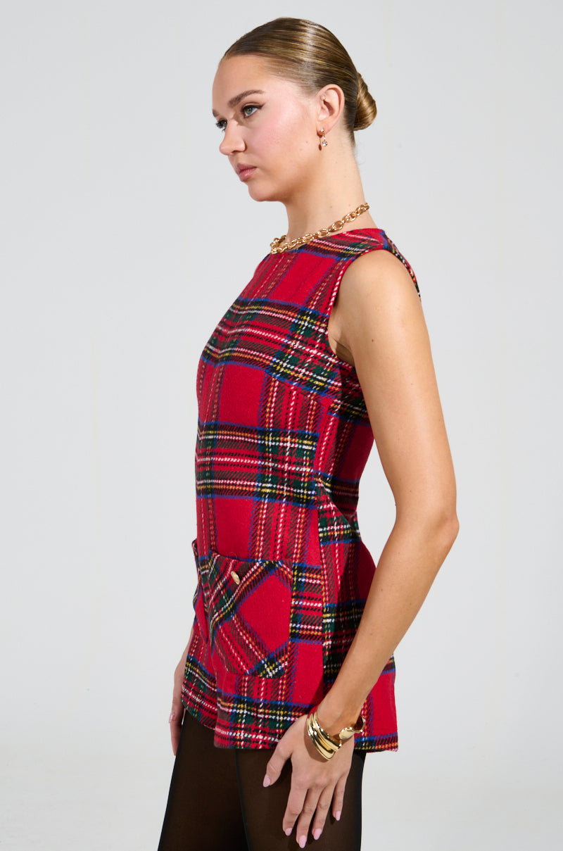 A woman wearing the Best Dressed Plaid Mini Dress in Red, featuring a sleeveless design and tweed-like fabrication, stands in profile against a plain background. She pairs it with black leggings and accessorizes with gold hoop earrings and bracelets, her hair styled in a sleek bun.