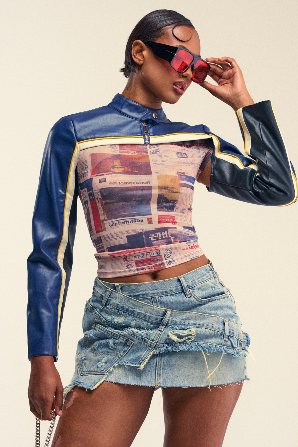 A stylish person wearing red sunglasses, a fitted top with graphic prints and the CAN I KICK IT BOLERO IN NAVY featuring blue faux leather moto sleeves, paired with a frayed denim mini skirt, poses confidently against a plain light background.