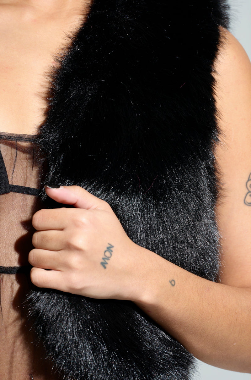 Dressed in the Lights Out Faux Fur Vest in black, a person sporting a sheer black top and a cropped sleeveless silhouette clasps the vest closed. A small tattoo adorns their hand, while another graces their upper arm against a neutral gray background.
