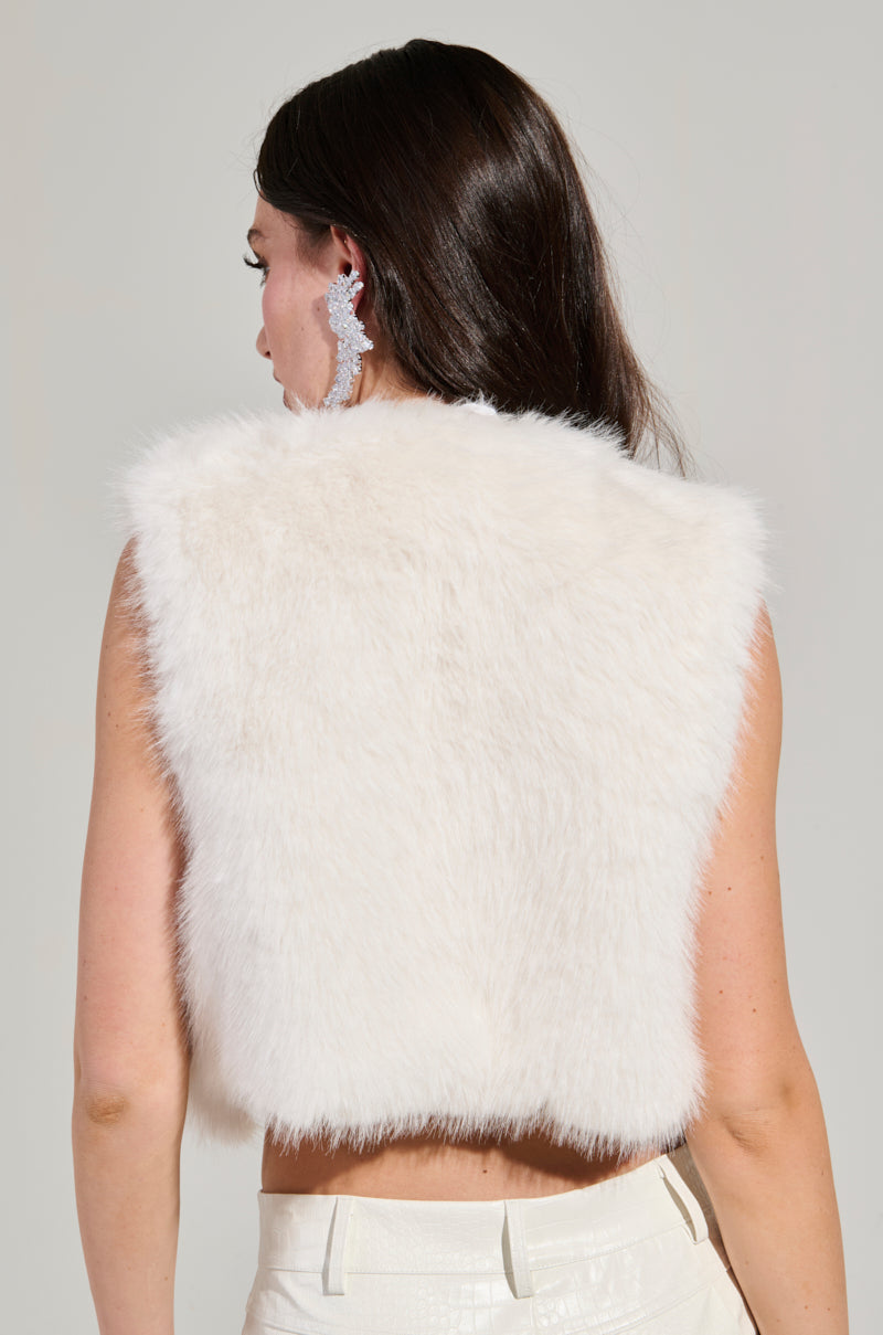 A woman with long dark hair is turned away, sporting the chic Lights Out Faux Fur Vest in white, lined with satin. She accessorizes with sparkly, dangling earrings and pairs it with light-colored pants against a plain, neutral background.