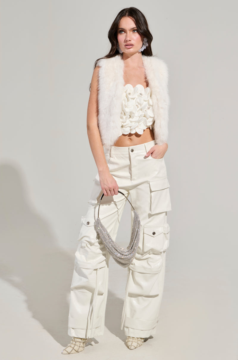 A person poses in a stylish outfit featuring the LIGHTS OUT FAUX FUR VEST IN WHITE, paired with a textured white crop top and cream cargo pants. They hold a sparkly handbag and wear pointed shoes, standing confidently against a plain background.