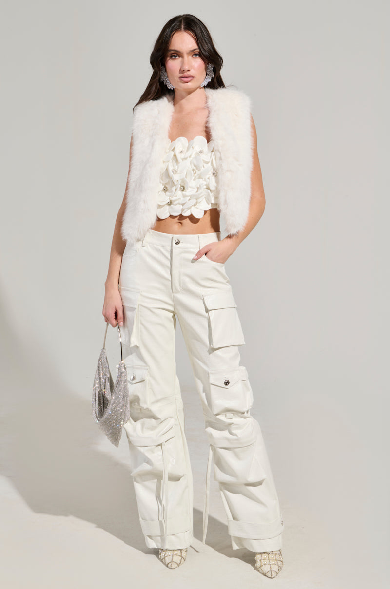 A woman poses in a stylish ensemble featuring the "Lights Out Faux Fur Vest in White," paired with a satin-lined, textured crop top and white cargo pants. She accessorizes with a sparkling silver handbag and statement earrings, while patterned shoes complete the look.