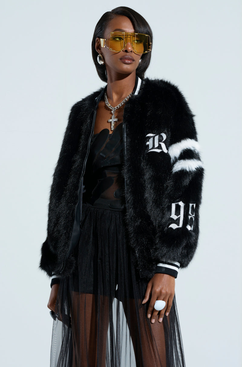 A person wearing a TRES CHIC FAUX FUR BOMBER featuring stylish white letters and numbers, paired with a sheer black skirt and yellow sunglasses. The look is complemented by hoop earrings, a cross necklace, and rings, making this cozy outerwear ensemble pop against the plain white background.