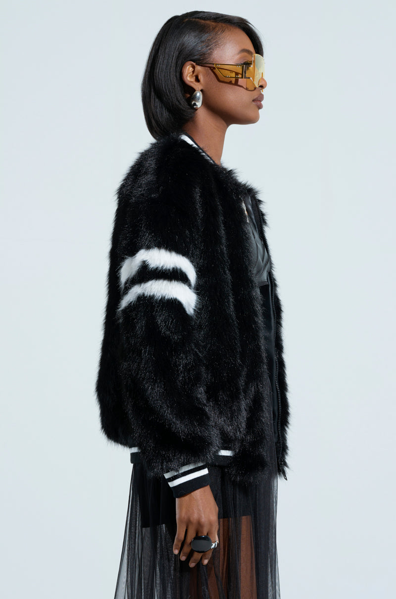 Profile of a woman showcasing the TRES CHIC FAUX FUR BOMBER, characterized by its black faux fur design and white stripes, complete with a ribbed knit collar. She accessorizes with yellow-tinted sunglasses and large earrings and features short, sleek hair and a black outfit underneath, all set against a plain background.