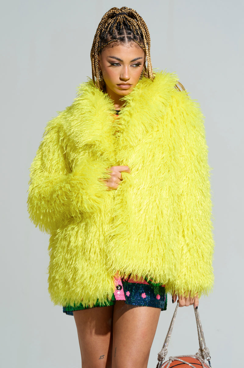 A person in the HIGHLIGHT OF YOUR NIGHT FAUX FUR JACKET, vibrant in bright yellow, paired with denim shorts and sporting braided hair, is holding a textured handbag embellished with rhinestone accents against a plain and neutral background.