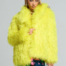 A person in the HIGHLIGHT OF YOUR NIGHT FAUX FUR JACKET, vibrant in bright yellow, paired with denim shorts and sporting braided hair, is holding a textured handbag embellished with rhinestone accents against a plain and neutral background.