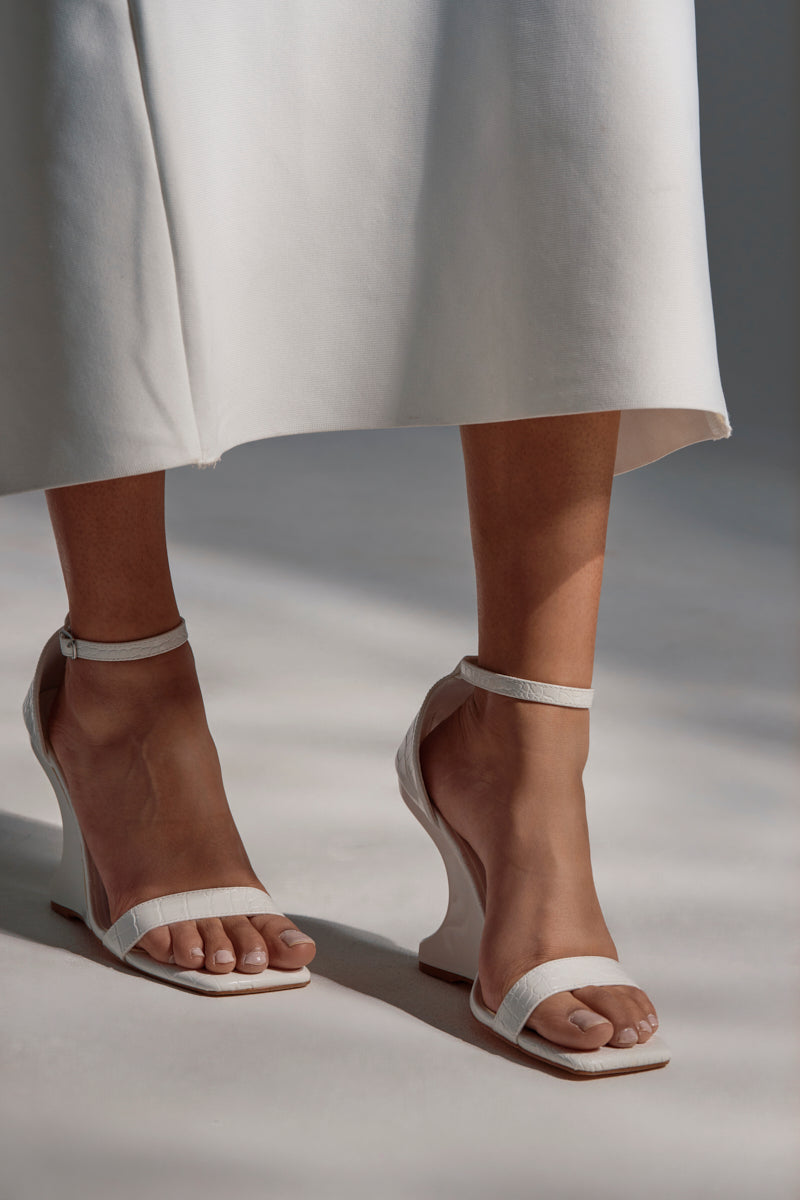 A person wearing a white midi skirt and chic AKIRA AALTO WHITE SANDAL wedges with ankle straps and inverted heels, standing on a neutral-colored floor.