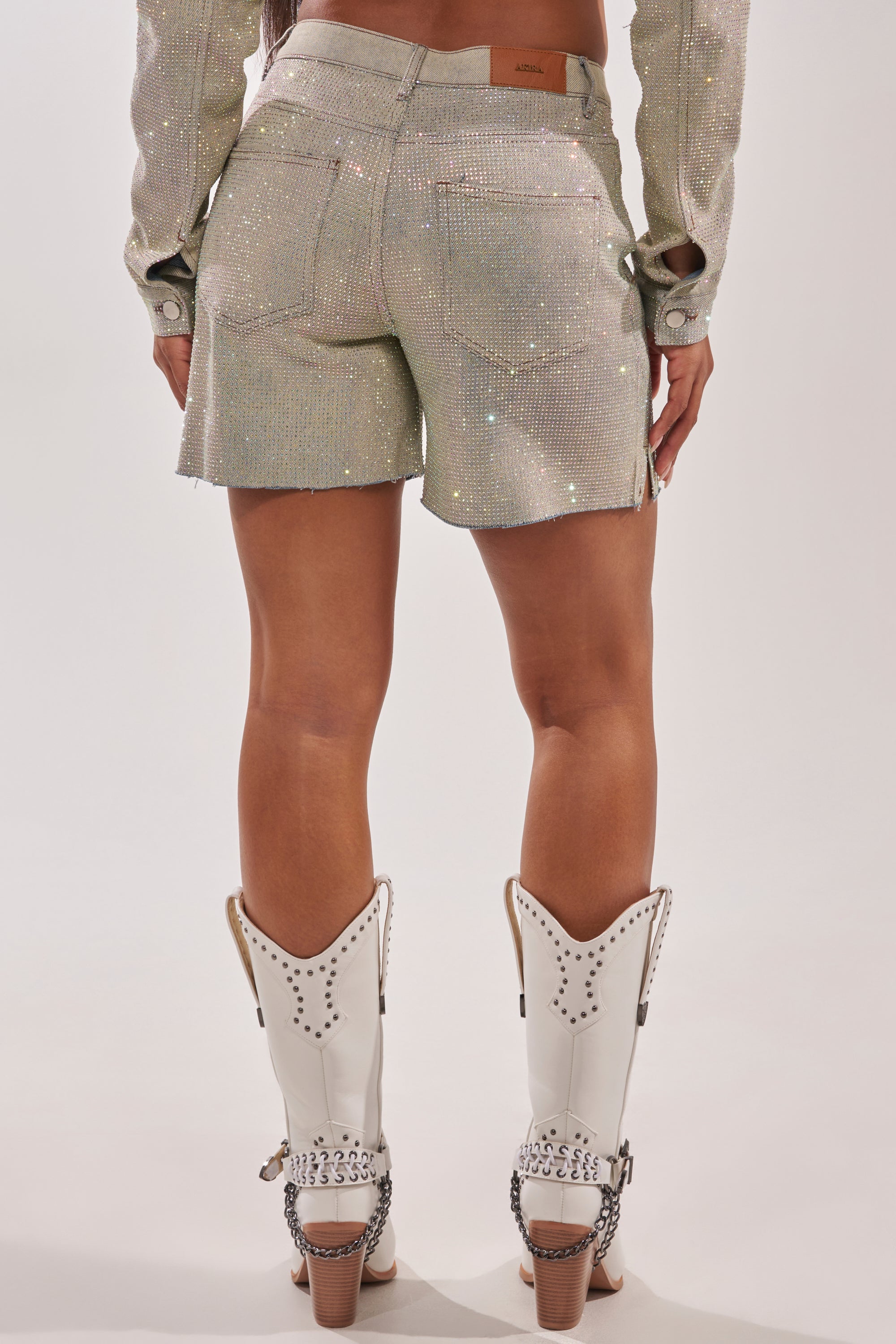 A person stands facing away in a well-lit studio, wearing SHINE BRIGHT SHORTS in sparkly silver, a matching long-sleeve top, and white cowboy boots with metal heel details. 10