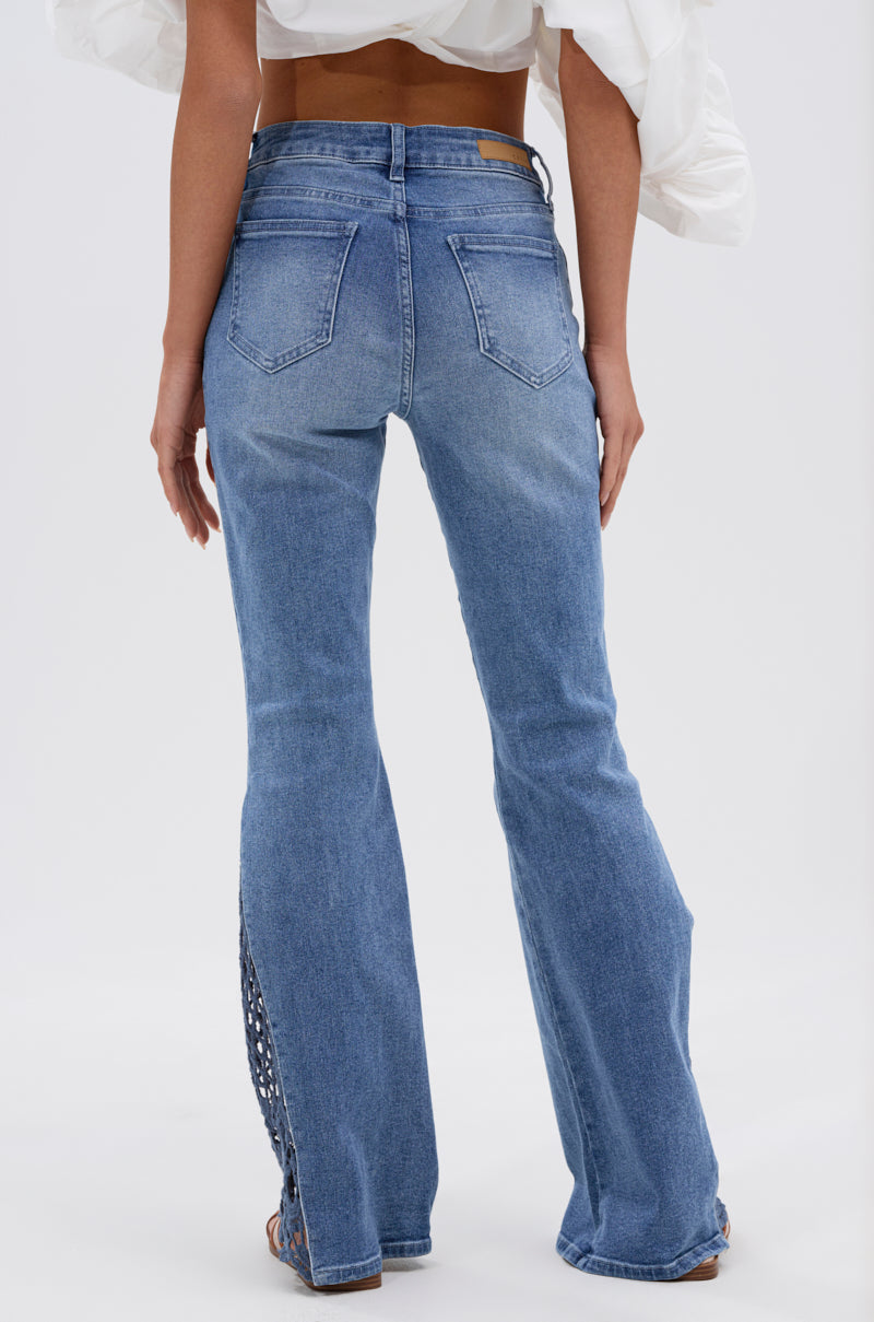 A woman is seen from the back wearing SUMMER DAYS RELAXED DENIM WITH EYELET OPENING—high-waisted blue flared jeans with back pockets and a lace-up eyelet detail on the lower leg, paired with a white off-shoulder puffy-sleeved top. 3