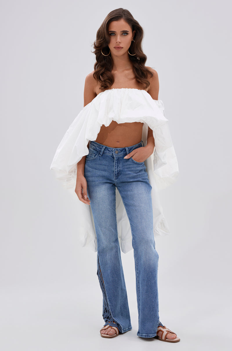 A woman poses against a plain background, wearing a white off-the-shoulder ruffled top and the SUMMER DAYS RELAXED DENIM WITH EYELET OPENING, paired with brown sandals. She has wavy brown hair and one hand in her pocket. 4