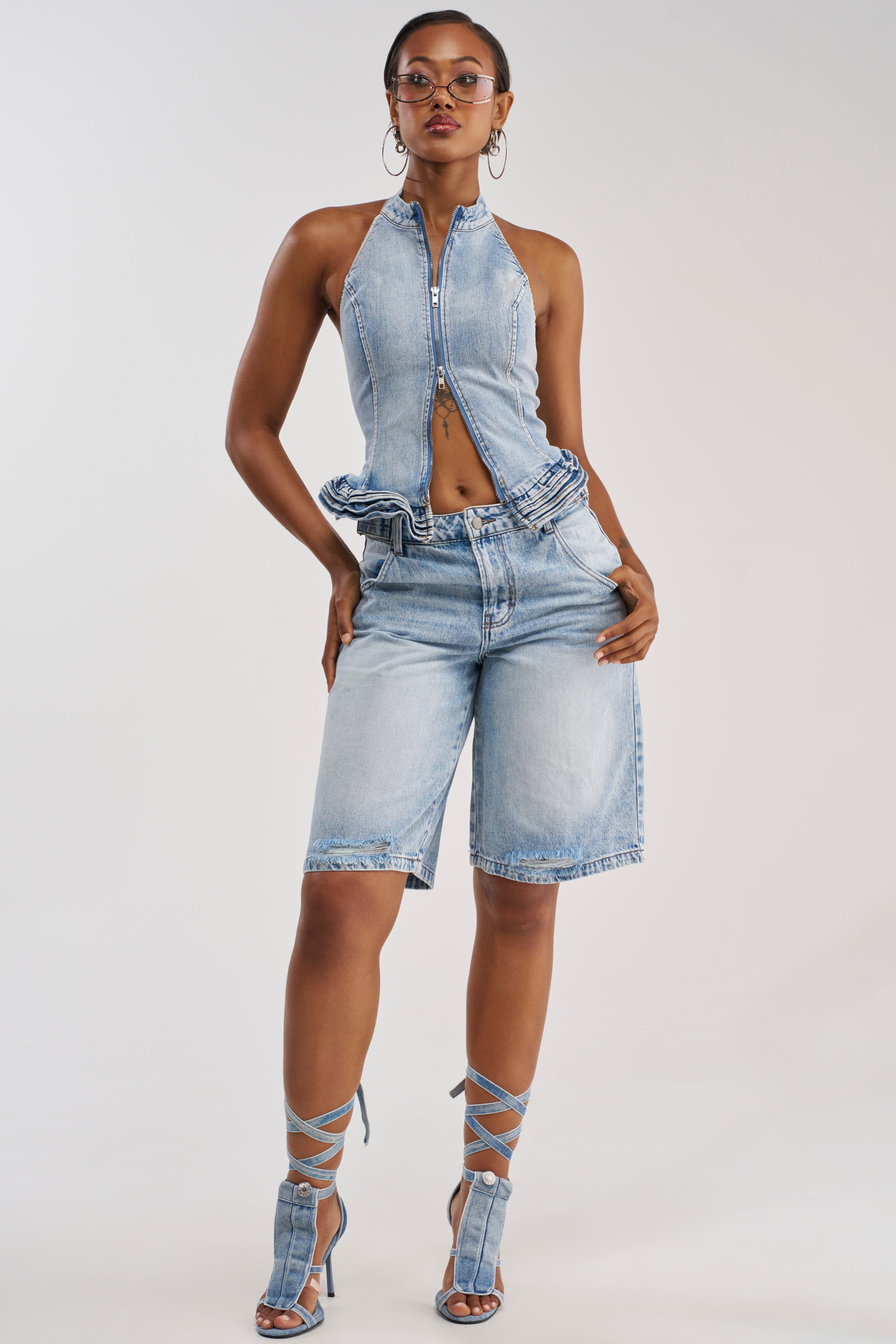 A woman poses in the FEEL GOOD DENIM SHORT paired with a light denim halter top, hoop earrings, and strappy high-heeled sandals for an effortlessly stylish summer streetwear look.