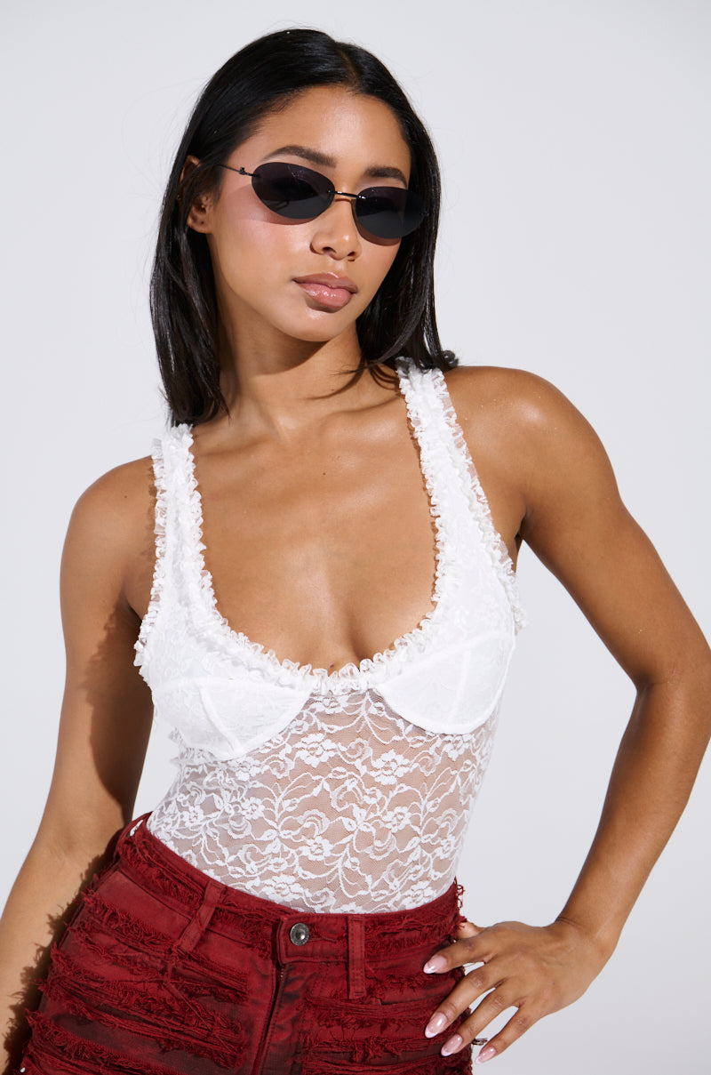 A woman confidently poses in the DAINTY LIL THING LACE SLEEVELESS BODYSUIT, featuring chic black sunglasses and striking red velvet shorts against a plain background.
