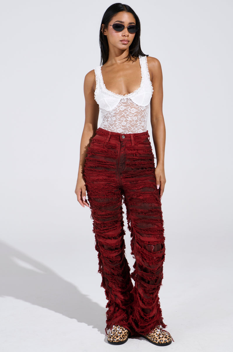 A woman confidently stands in a floral DAINTY LIL THING LACE SLEEVELESS BODYSUIT and distressed red pants. She wears sunglasses and animal print shoes, with the sheer fabric adding allure. The background is plain white.