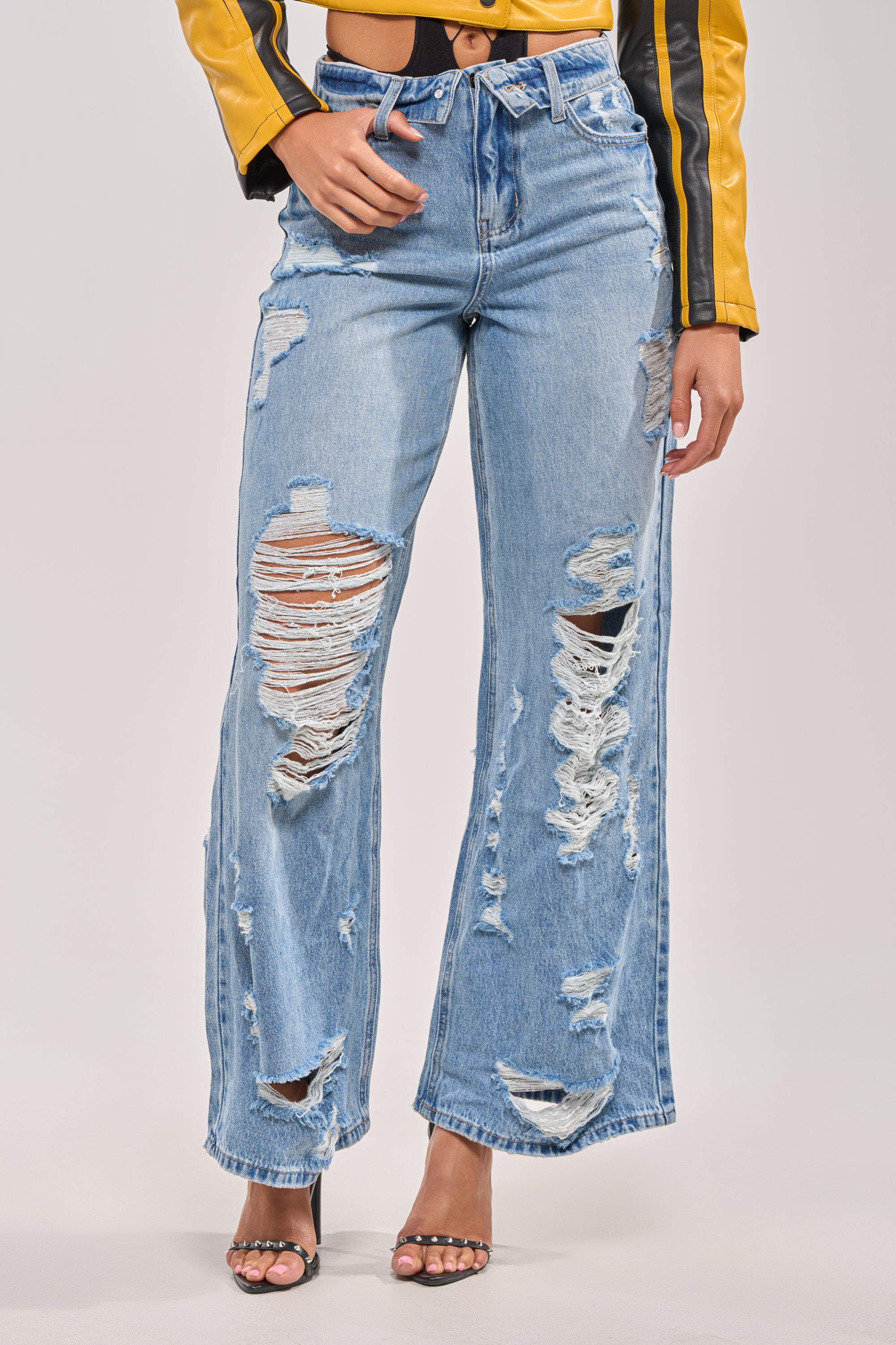 A person stands against a plain background wearing the SOMETHING NEW FOLD OVER WAISTBAND DISTRESSED DENIM jeans, styled with a yellow jacket featuring black stripes and black heeled sandals. 0