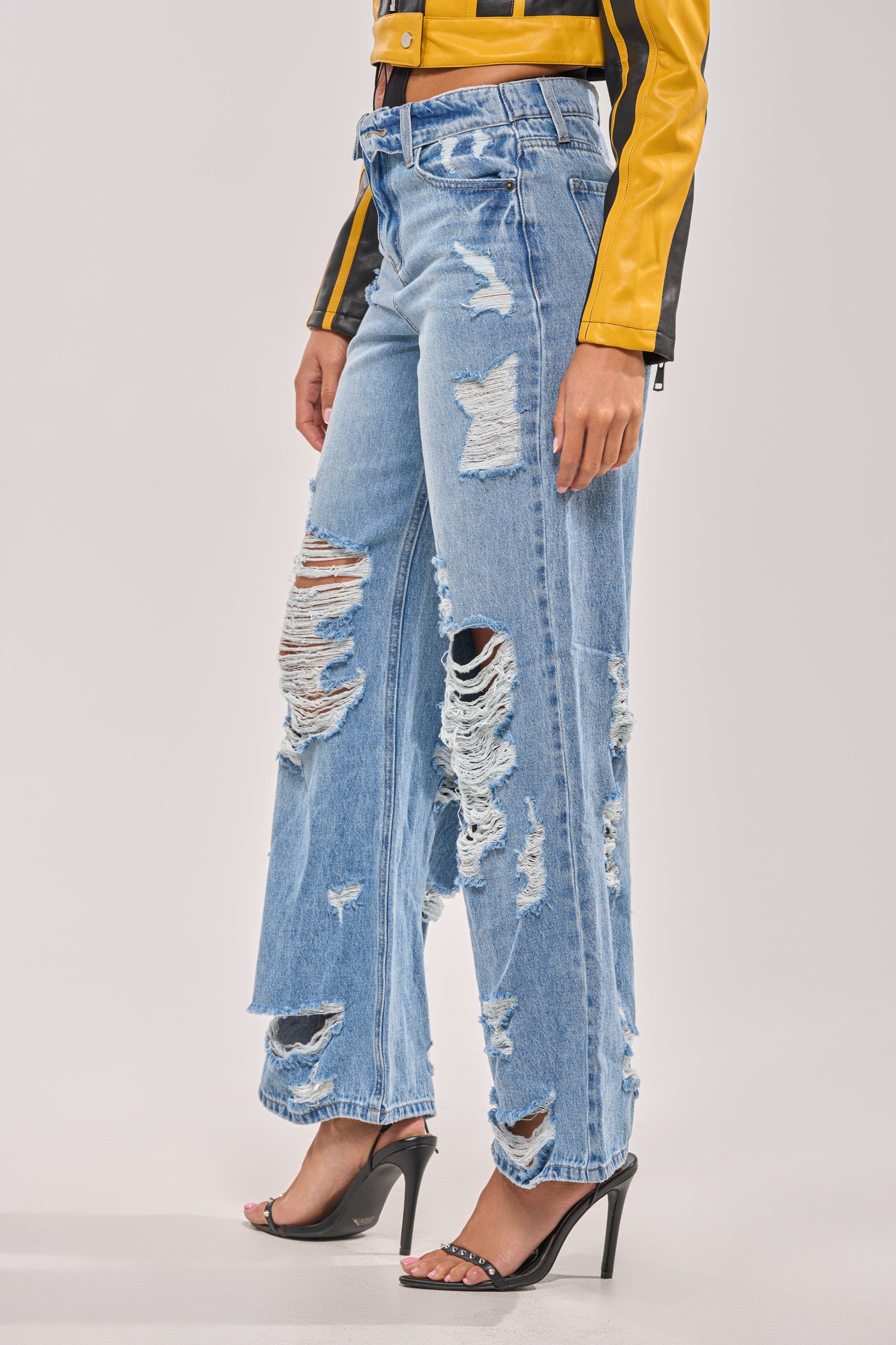 A person models the SOMETHING NEW FOLD OVER WAISTBAND DISTRESSED DENIM, paired with a yellow and black jacket and black high-heeled sandals against a plain background. 2