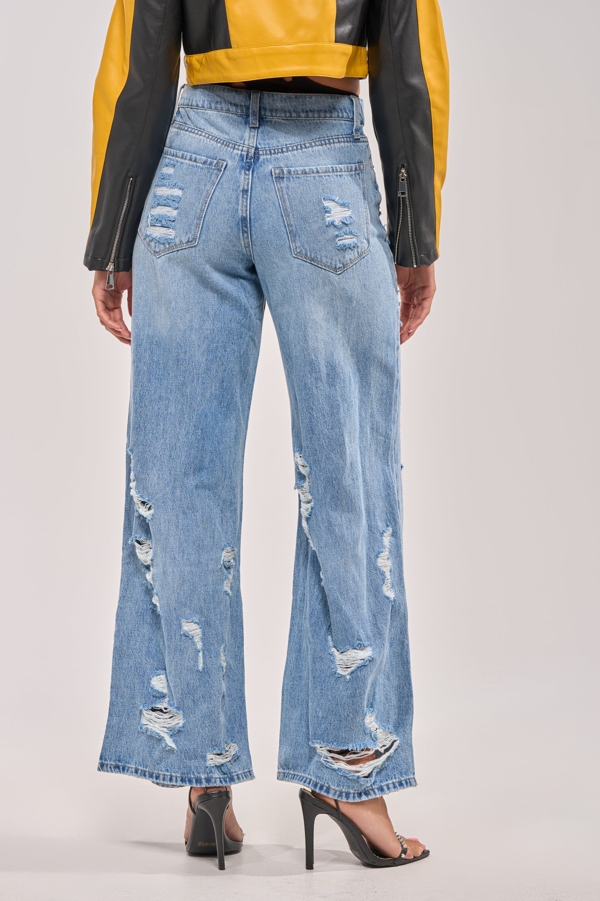 A person is shown from the back wearing a yellow and black jacket, SOMETHING NEW FOLD OVER WAISTBAND DISTRESSED DENIM jeans in a light wash, and black high-heeled sandals against a plain background. 3