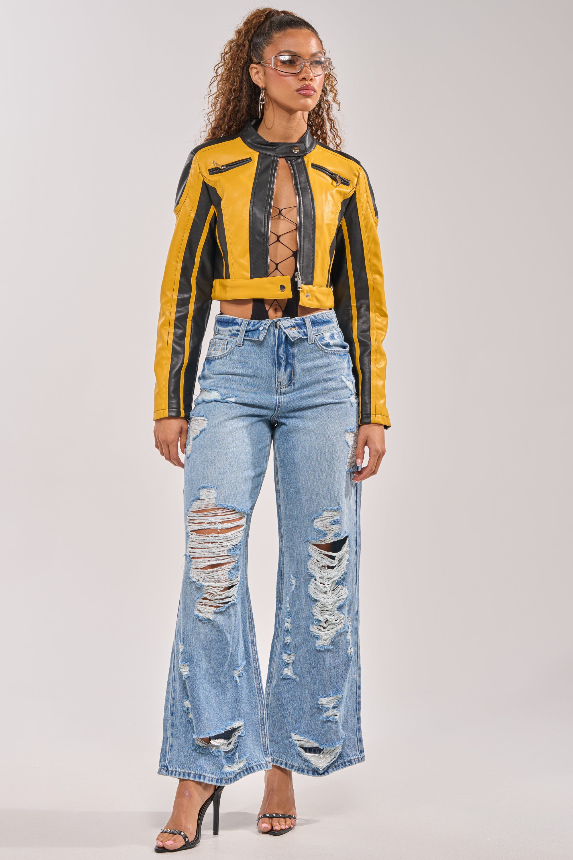 A woman with curly hair and clear-framed glasses wears a yellow and black cropped leather jacket, a lace-up black top, SOMETHING NEW FOLD OVER WAISTBAND DISTRESSED DENIM, and black high-heeled sandals. 4