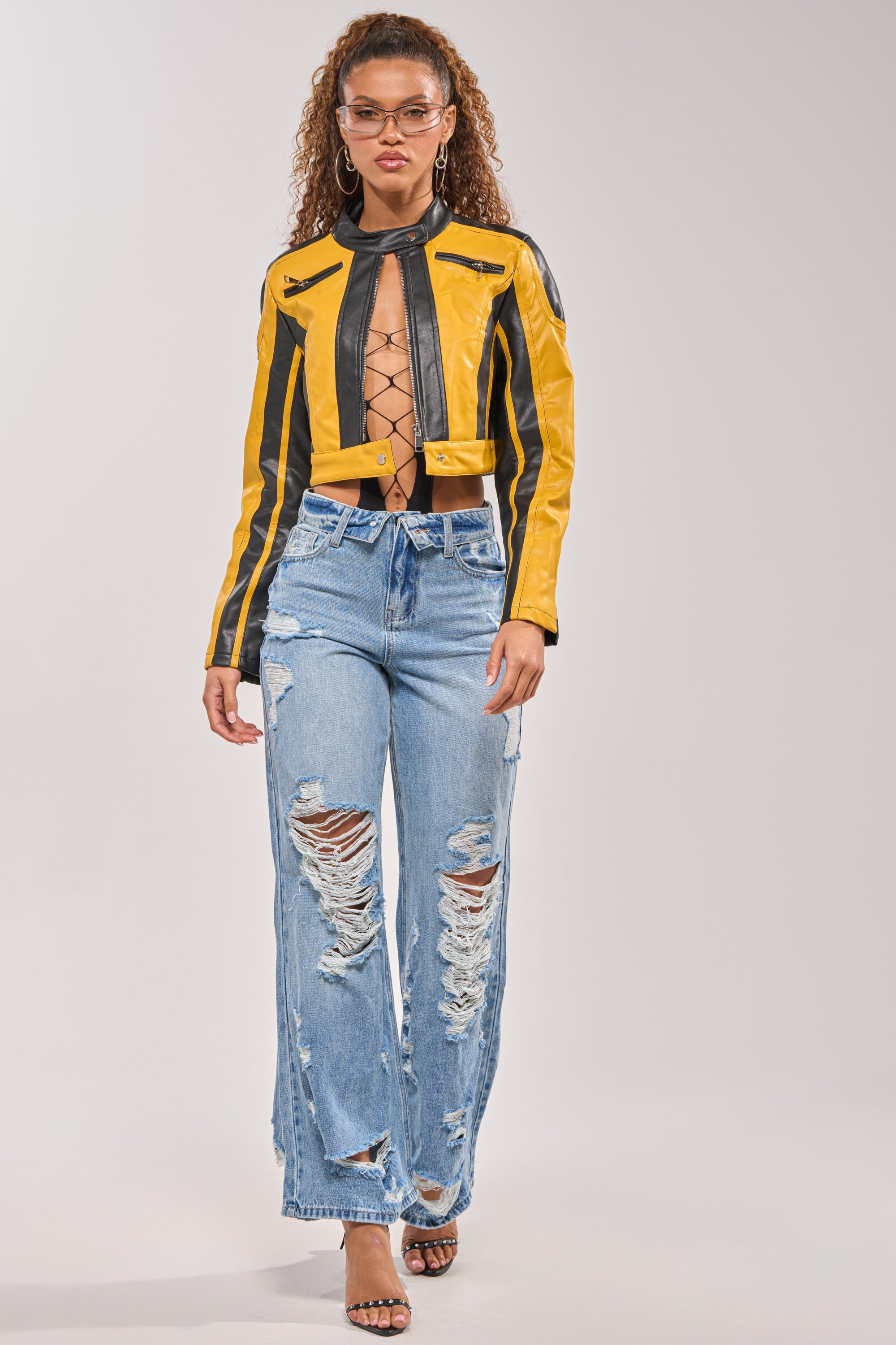 A person with curly hair confidently walks against a plain background, wearing a yellow and black cropped leather jacket, a lace-up black top, strappy heels, and the SOMETHING NEW FOLD OVER WAISTBAND DISTRESSED DENIM. 5