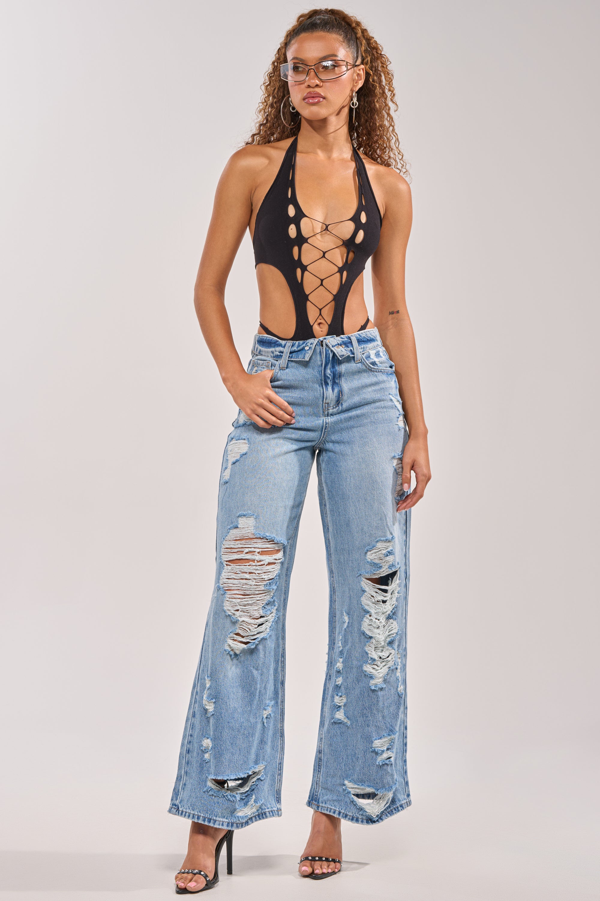 A woman models the SOMETHING NEW FOLD OVER WAISTBAND DISTRESSED DENIM with a black lace-up cutout bodysuit, clear heels, and transparent glasses, posing confidently against a plain background. 6