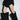 A person in a short black dress with ruffled layers, standing indoors against a white wall, wears the AZALEA WANG ABAWI BLACK EXTREME BOOT IN SUEDE—striking pointed-toe boots that reach above the knees.