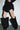 A person in a short black dress with ruffled layers, standing indoors against a white wall, wears the AZALEA WANG ABAWI BLACK EXTREME BOOT IN SUEDE—striking pointed-toe boots that reach above the knees.