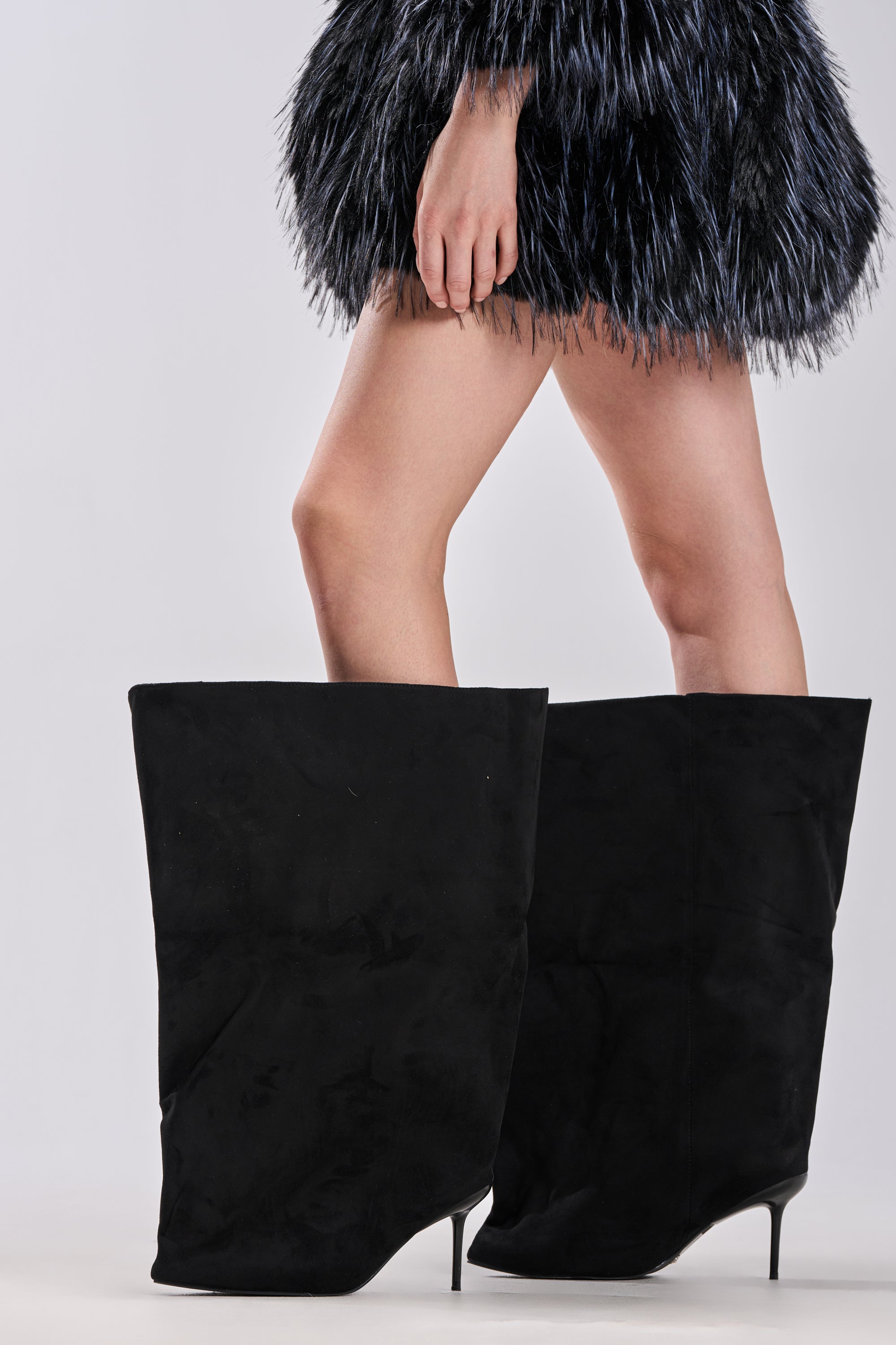 A person wearing an oversized black furry dress and the AZALEA WANG ABAWI BLACK EXTREME BOOT IN SUEDE with a pointed toe stands against a plain light gray background. 1