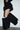 A person, shown from the waist down, stands against a plain white wall wearing a short, ruffled black skirt and AZALEA WANG ABAWI BLACK EXTREME BOOT IN SUEDE stilettos.