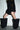 Wearing a black ruffled mini skirt, black long-sleeve top, and AZALEA WANG ABAWI BLACK EXTREME BOOT IN SUEDE, a person stands against a white wall with their face out of frame.