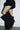 Wearing black ruffled shorts and the AZALEA WANG ABAWI BLACK EXTREME BOOT IN SUEDE—knee-high, oversized stiletto heel boots—a person stands against a white wall with their legs crossed.
