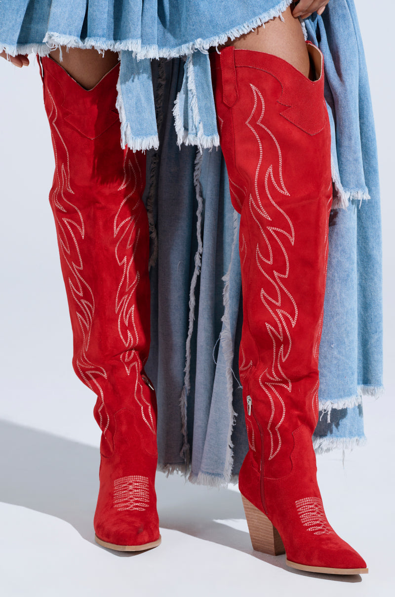 A person wears the ABBY RED HIGH SHAFT WESTERN BOOT, characterized by flame-inspired embroidery and a slanted wooden heel, paired with a light blue frayed denim skirt. 0