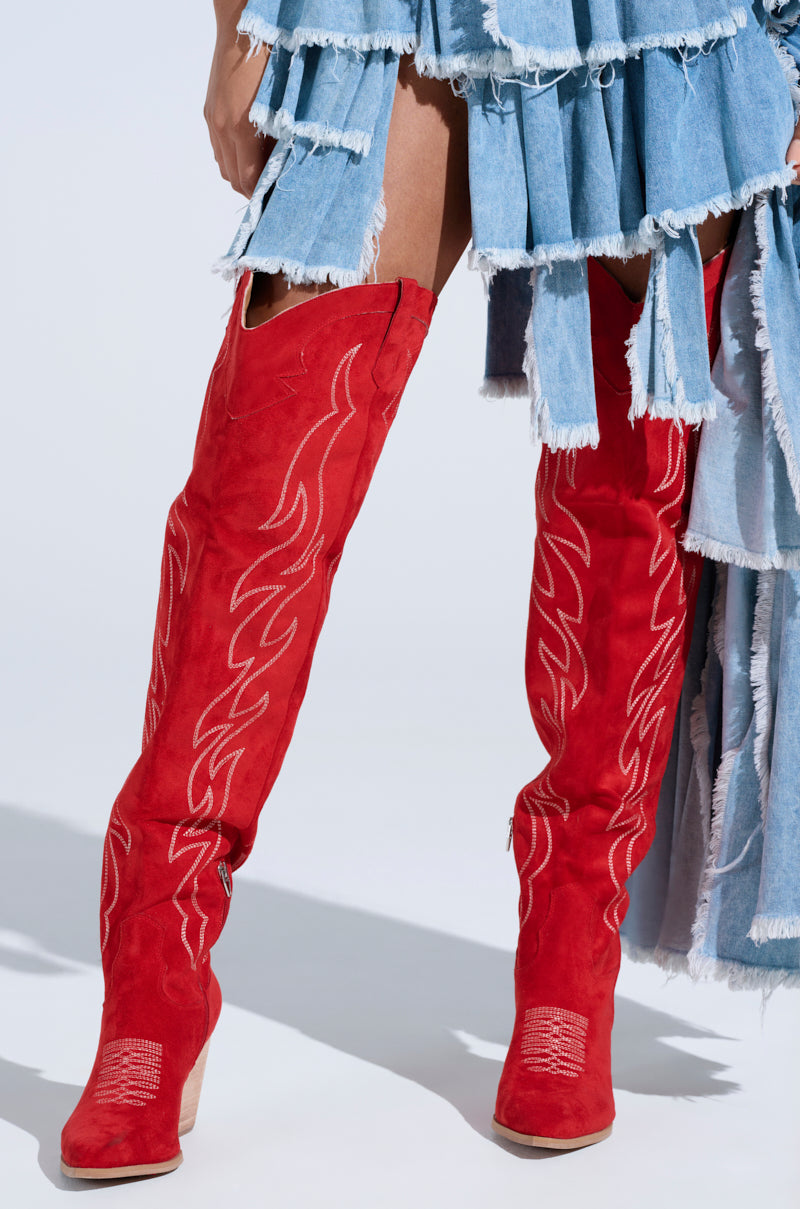 Someone wears an ABBY Red High Shaft Western Boot in faux suede with flame-inspired embroidery, paired with a frayed denim skirt. The boot's decorative stitching contrasts with the skirt's ragged edges, all set against a plain white backdrop. 1
