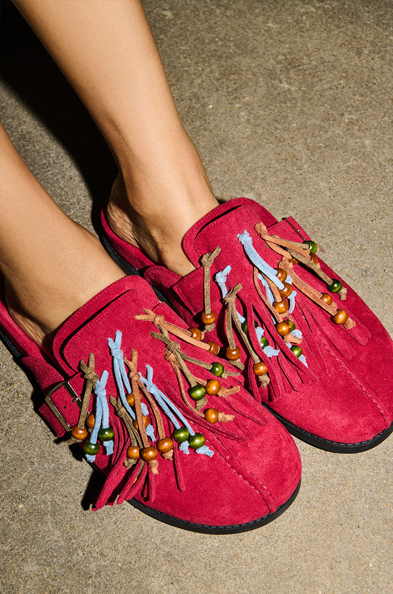 Person wearing the AZALEA WANG ABEILLE BURGUNDY FAUX SUEDE FRINGE CLOG, featuring bright fringe and a bold look, stands on a textured concrete surface.