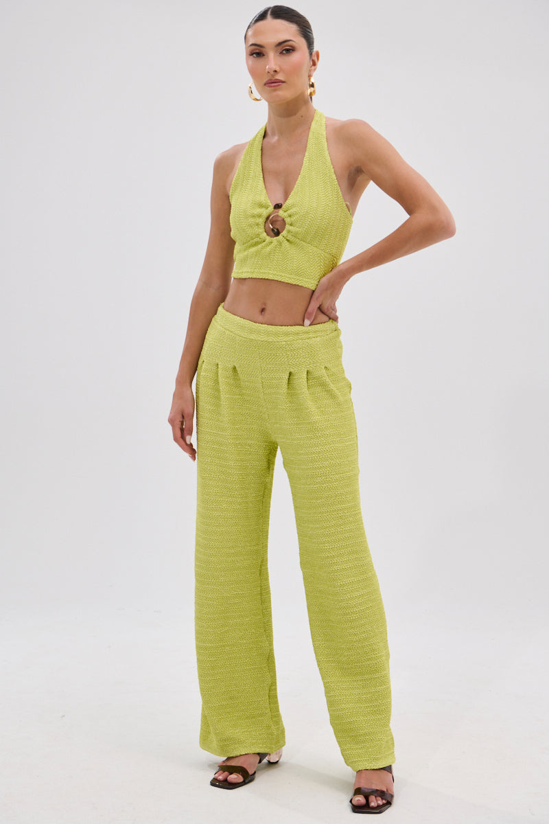 A woman wears the HOLLYWOOD SLINKY TEXTURED PALAZZO PANT IN GREEN with a matching light green knit halter crop top featuring a ring detail. Her hair is pulled back, and she accessorizes with sandals and statement earrings against a plain background.