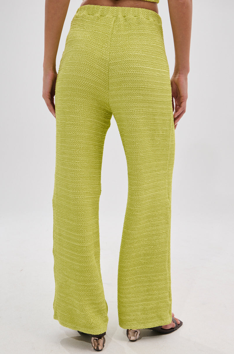 A person faces away, wearing the HOLLYWOOD SLINKY TEXTURED PALAZZO PANT IN GREEN with a high-rise fit, slight flare, and open-toe heels, photographed against a plain white background.
