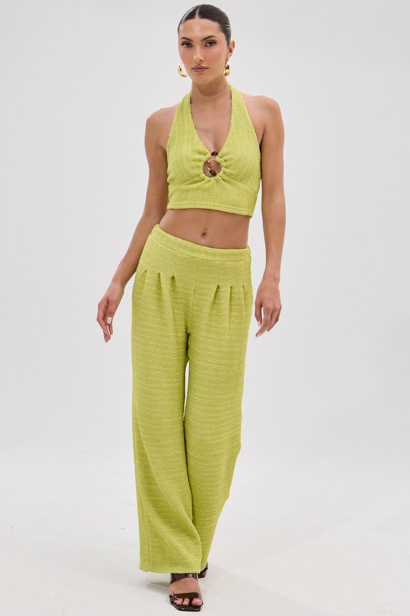 A woman poses in HOLLYWOOD SLINKY TEXTURED PALAZZO PANT IN GREEN with a matching light green halter crop top, ring chest detail, elastic waistband, and black heeled sandals against a plain white background.