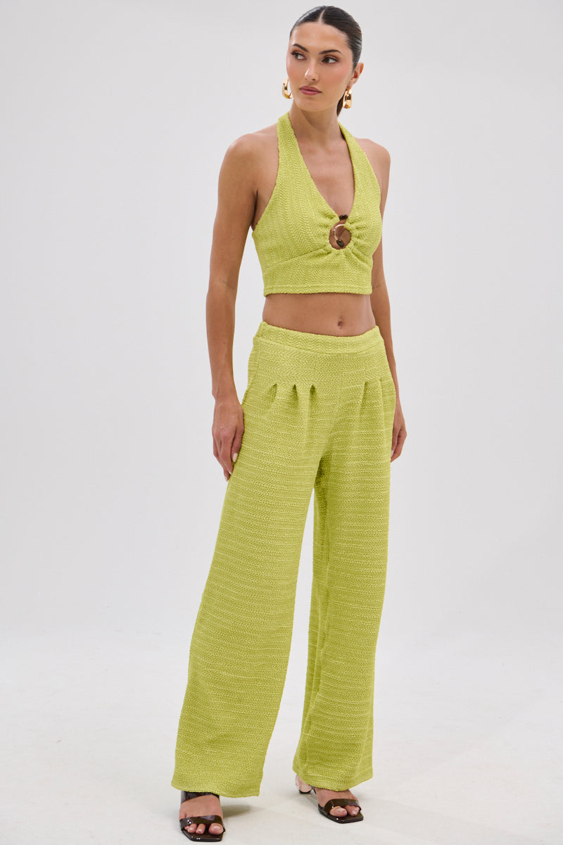 A woman wears the HOLLYWOOD SLINKY TEXTURED PALAZZO PANT IN GREEN with a matching light green halter crop top featuring a ring detail, standing against a white background styled with hoop earrings and heels.